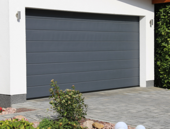 The image shows a closed, modern garage door. The garage door is gray and segmented horizontally. The surrounding area includes a paved driveway with a pattern of rectangular stones, and there are some plants and small shrubs in a landscaped area adjacent to the driveway. The exterior wall of the garage is white, and there are two wall-mounted lights on either side of the garage door. Associated with Pride Garage Door.