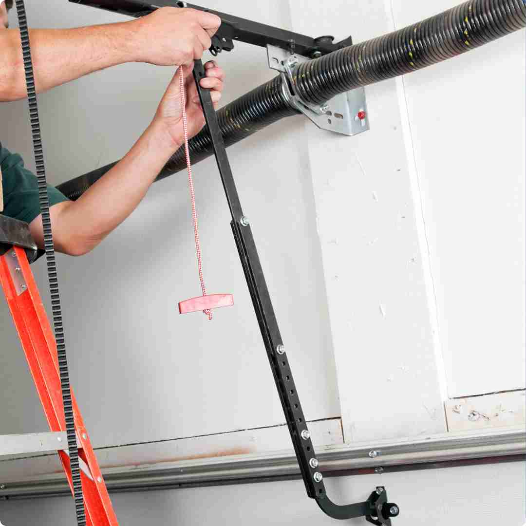 Close-up view of a garage door mechanism, showing a torsion spring mounted on a metal rod, brackets securing the rod to the ceiling, and a black metal bar connected to the garage door opener mechanism. A person is holding a tool and a red and white rope, likely adjusting or repairing the garage door system. Associated with Pride Garage Door.