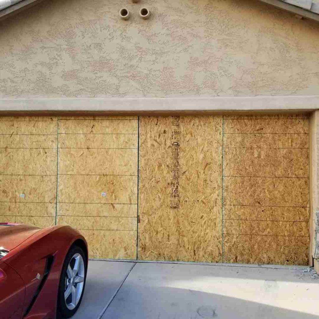The image shows a garage door that has been boarded up with plywood sheets. The garage door appears to be a double door, and the plywood sheets cover the entire surface. Two circular vents are above the garage door on the wall, and a red car is partially visible in the lower left corner, parked in front of the garage. The plywood sheets have some markings and lines on them, possibly indicating measurements or cuts. Associated with Pride Garage Door.