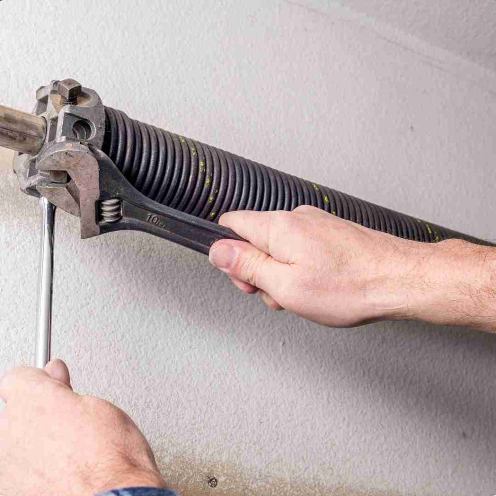 Close-up view of a garage door mechanism, showing a torsion spring mounted on a metal rod, brackets securing the rod to the ceiling, and a black metal bar connected to the garage door opener mechanism. A person is holding a tool and a red and white rope, likely adjusting or repairing the garage door system. Associated with Pride Garage Door.