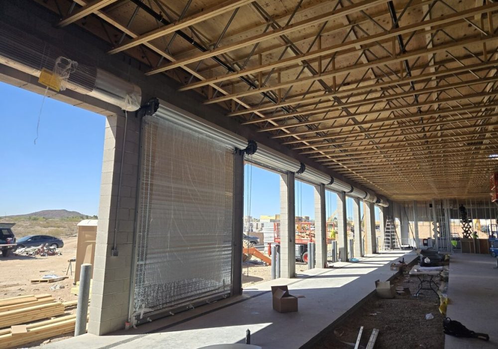 Interior of a construction site with exposed wooden beams, large roll-up doors, and scattered building materials, under a clear blue sky.