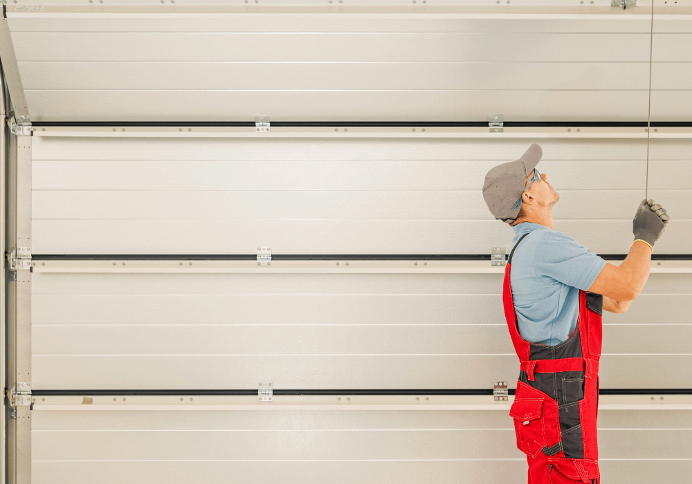 A man in red overalls and gray cap pulls a garage door release cord, with the door visible above him in a brightly lit garage.