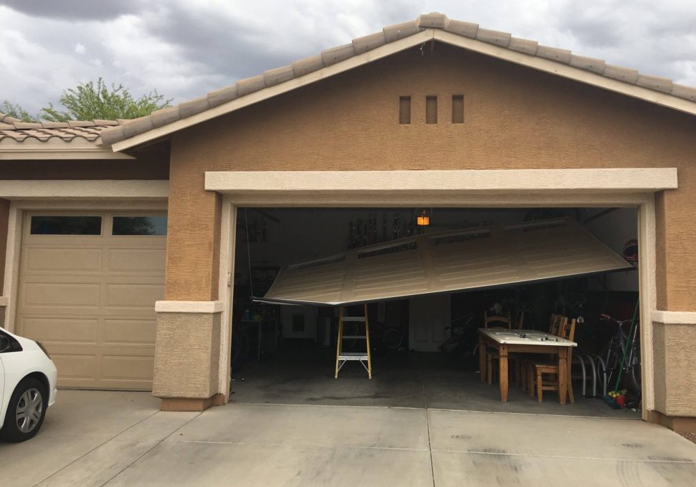The image shows a garage with a partially broken garage door. The garage door is tilted and appears to be off its tracks, with the left side hanging lower than the right side. Inside the garage, there is a yellow step ladder positioned under the garage door, possibly indicating an attempt to fix it. There is also a wooden table with chairs and various items scattered around the garage. To the left of the broken garage door, there is another closed garage door, and a white car is partially visible in the driveway. The sky above is cloudy, suggesting overcast weather. Associated with Pride Garage Door.