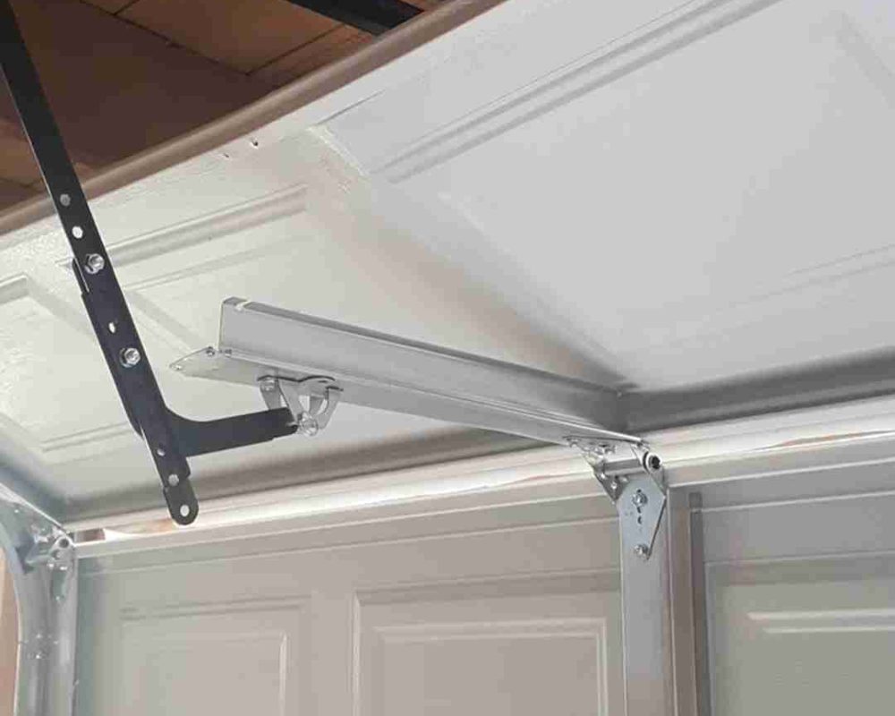 "Close-up view of the mechanism of a garage door opener. The garage door is partially open, and the image focuses on the metal track and the arm that connects the door to the opener. The metal track is mounted to the ceiling, and the arm is attached to the door with bolts. Associated with Pride Garage Door.
