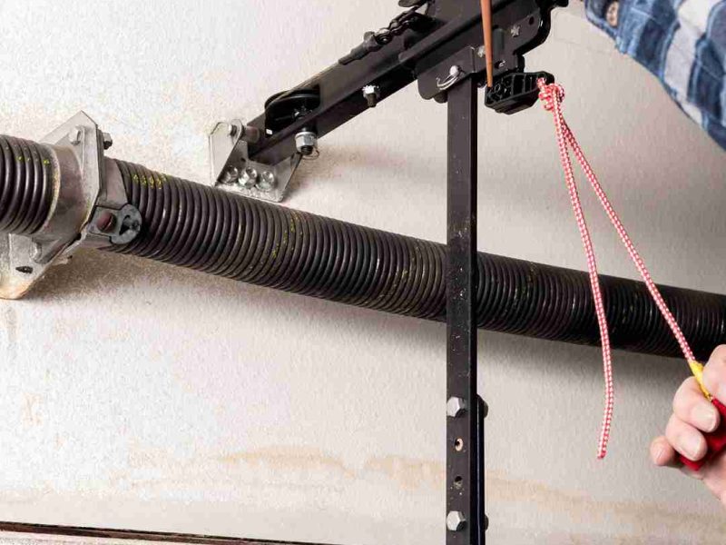 Close-up view of a garage door mechanism, showing a torsion spring mounted on a metal rod, brackets securing the rod to the ceiling, and a black metal bar connected to the garage door opener mechanism. A person is holding a tool and a red and white rope, likely adjusting or repairing the garage door system. Associated with Pride Garage Door.